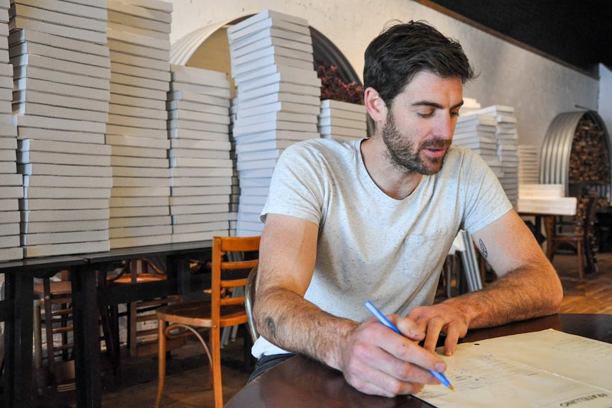 A man sits at a table with a pen and a menu, pizza boxes are piled up behind him.