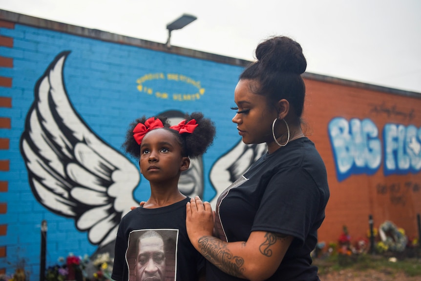 A black woman holds her arms around a little black girl with red ribbons in her hair in front of a mural of George Floyd