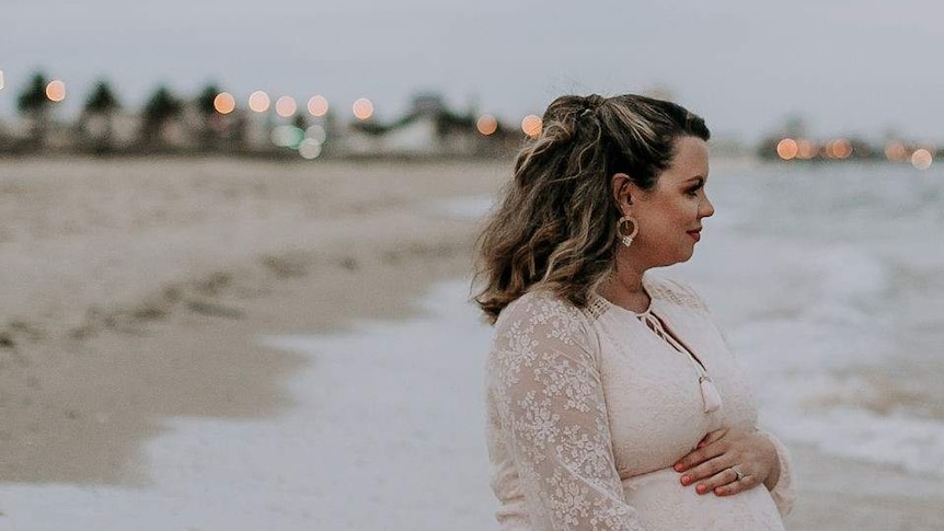 Leticia who is nearing the end of her pregnancy, sits on a beach wearing a beautiful pale pink lace dress while waves lap at her