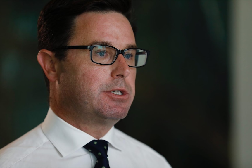 A middle-aged man with dark brown hair & dark-rimmed glasses looks frustrated as he speaks 