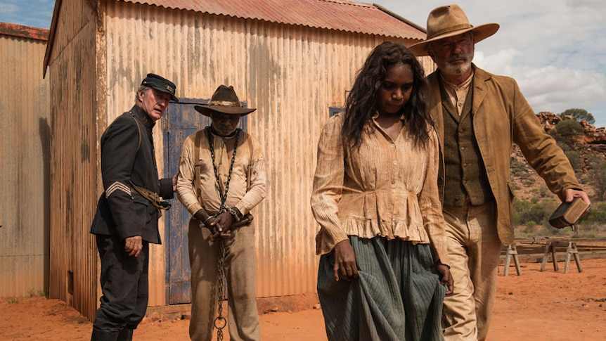 The four actors stand in tattered period clothing, one of them in chains, on a red dirt road in front of a corrugated iron shed.