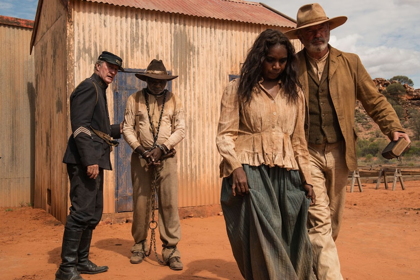 The four actors stand in tattered period clothing, one of them in chains, on a red dirt road in front of a corrugated iron shed.
