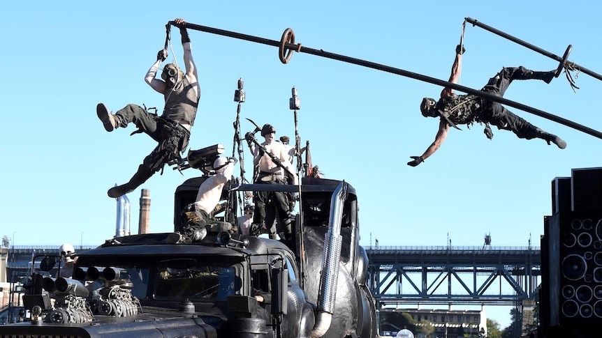 Cast members from 'Mad Max - Fury Road' swing on a vehicle from the movie during the filming a promo on the Sydney Opera House on May 13, 2015.