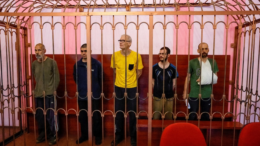 Five men stand in a cage in a courtroom.