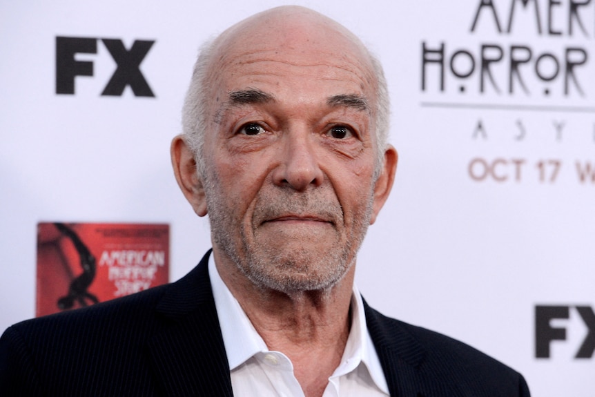 An elderly male actor on a red carpet, looks expressionless towards photographers