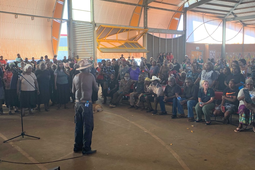 A crowd of Warlpiri Aboriginal people gathered and listening to a man with a microphone in the basketball community hall