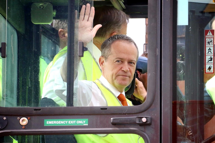 Bill Shorten waves to the media from inside the glass cab of large forklift.