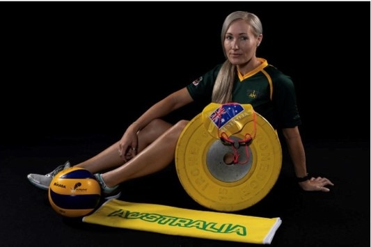 A woman with blonde hair sits down. There is a weight in front of her, and a ball at her foot.