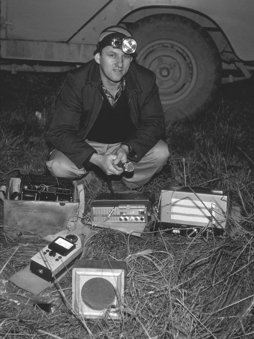 A black and white photograph of Murray Littlejohn holding a microphone as he crouches down with recording equipment at his feet.