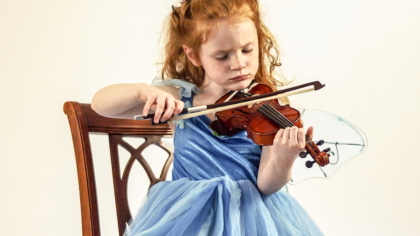 A child plays violin while sitting on a wooden chair and wearing a blue fairy dress.
