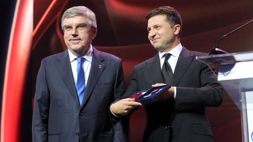 Ukrainian President Volodymyr Zelenskyy presents a medal to IOC president Thomas Bach on stage next to a lectern.