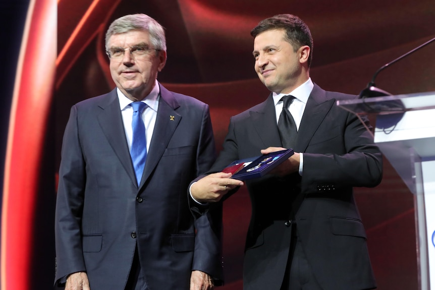 Ukrainian President Volodymyr Zelenskyy presents a medal to IOC president Thomas Bach on stage next to a lectern.