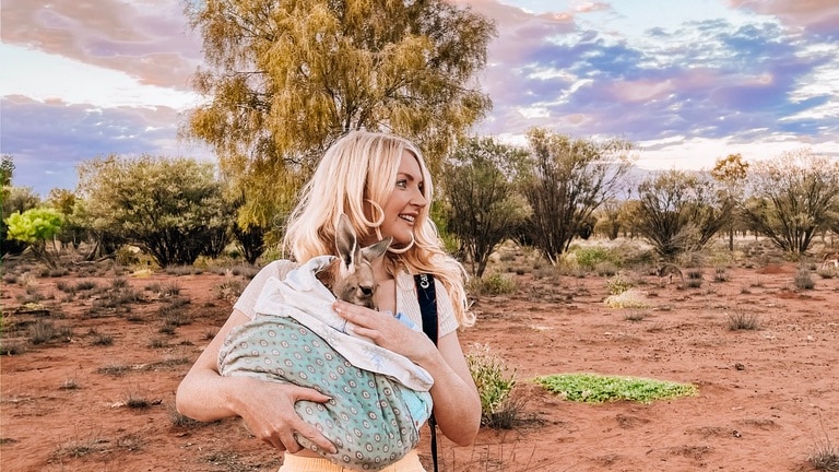 A woman in peach pants cuddles a small animal wrapped in a blanket in a red sandy desert