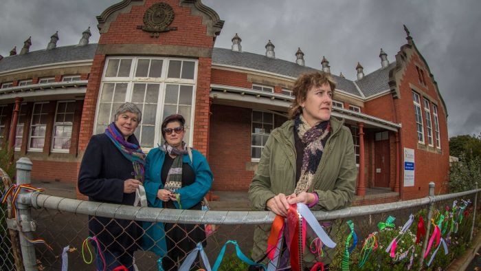 Three women stand behind a school fence with colourful ribbons tied to it.