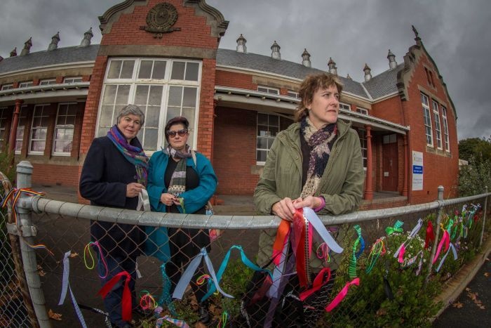 Three women stand behind a school fence with colourful ribbons tied to it.