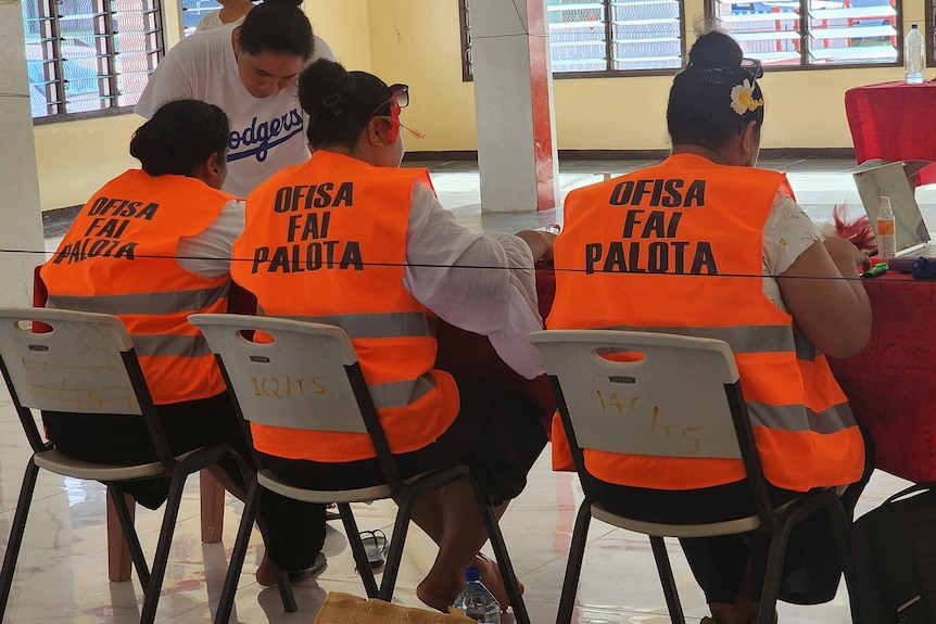 Electoral staff wearing neon orange bibs sit at desk counting votes.