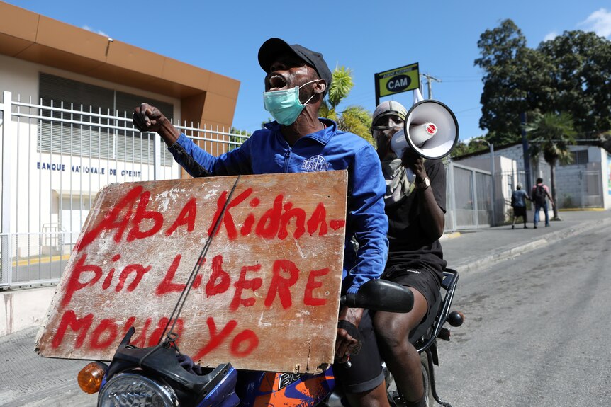 Two people on a motorcycle, the one in front holding a sign with French text, the one behind shouting into a megaphone