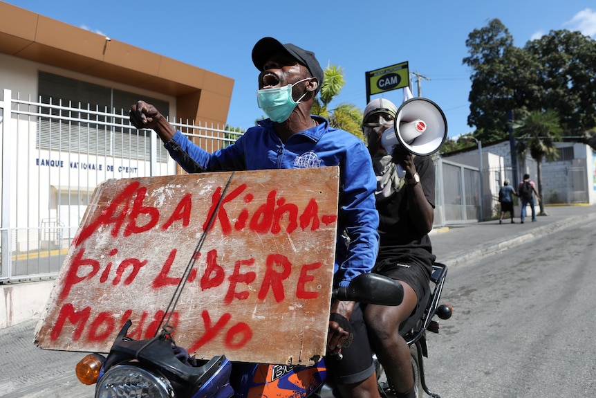 Two people on a motorcycle, the one in front holding a sign with French text, the one behind shouting into a megaphone