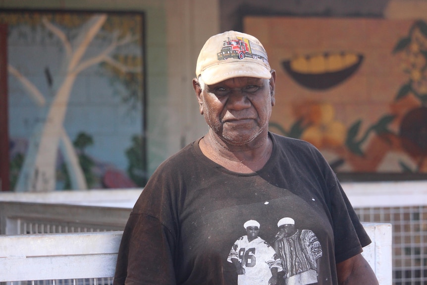 He wears a cap and stands by the Docker River store.