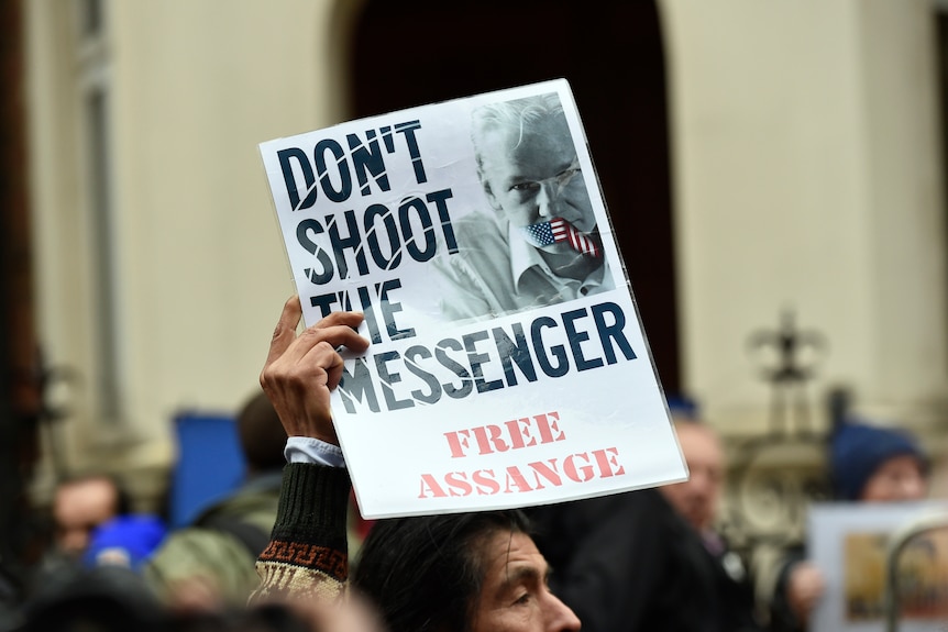 A man holds up a sign that reads Don't Shoot the Messenger, Free Assange with Julian's mouth taped over with the US flag
