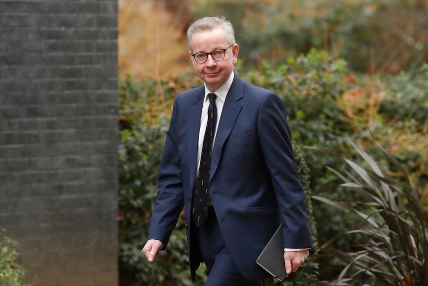 Michael Gove wearing glasses and a suit walks while holding a notebook.