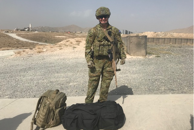 Une femme vêtue d'un camouflage militaire complet se tient debout et pose devant la caméra, avec un paysage aride et aride de l'Afghanistan derrière elle. 