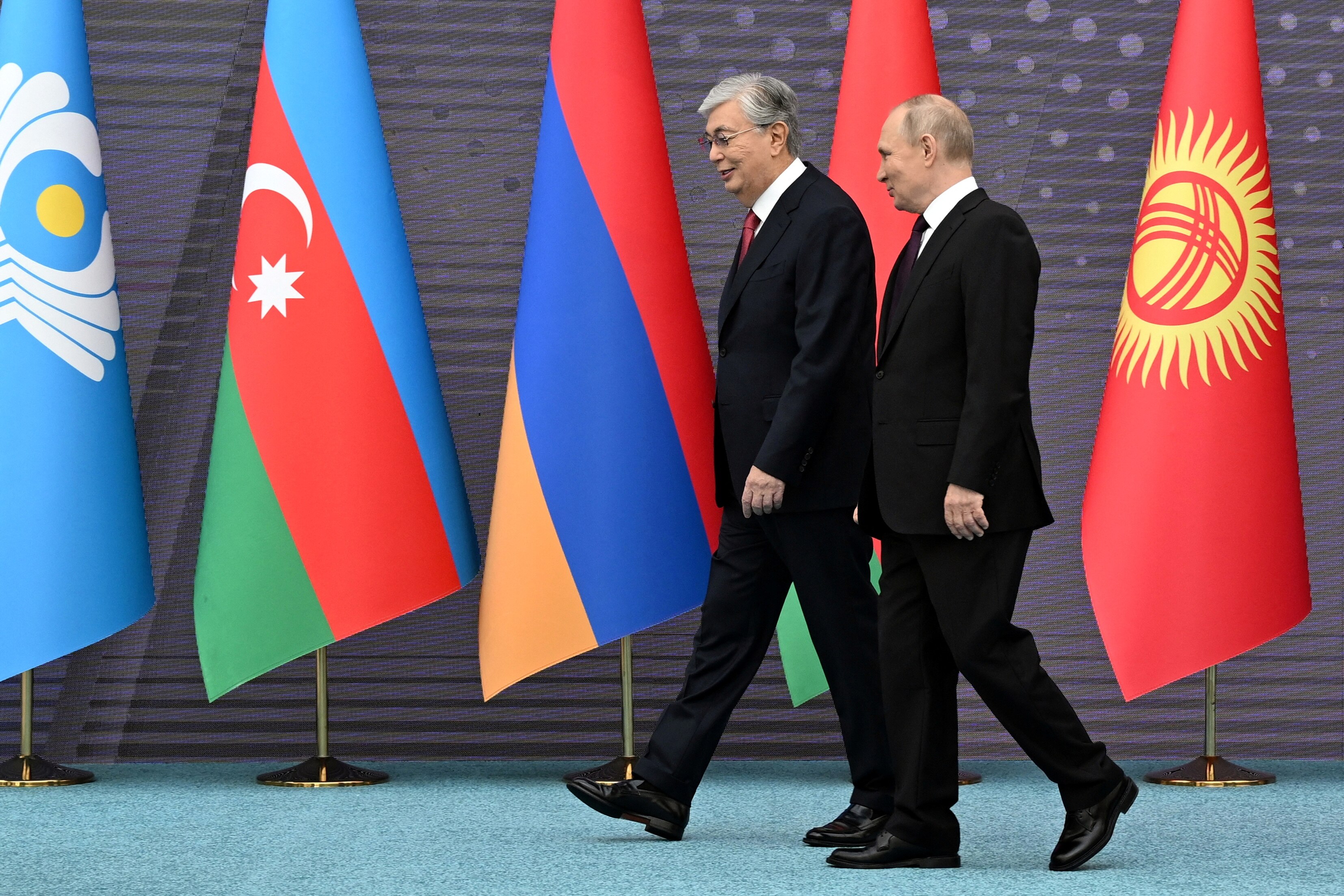 Vladimir Putin  leans his head towards a man with grey hair wearing a dark suit as they walk infront of flags.