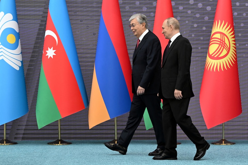 Vladimir Putin  leans his head towards a man with grey hair wearing a dark suit as they walk infront of flags.