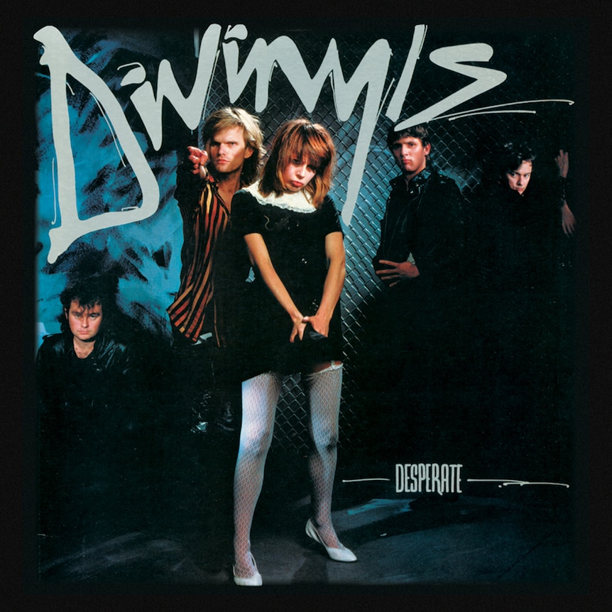 Five members of Divinyls pose in a dark alleyway in front of a chair fence. Chrissy Amphlett pouts at the camera