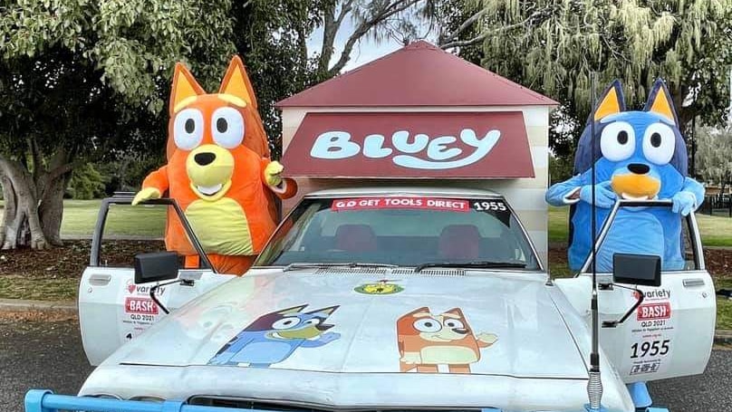 An image of a ute decorated with Bluey memorabilia and two adults in Bluey costumes standing next to two cars