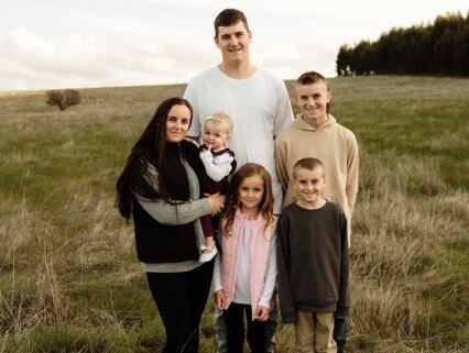 A woman holds a toddler while standing in a paddock with a tall man and three other children.