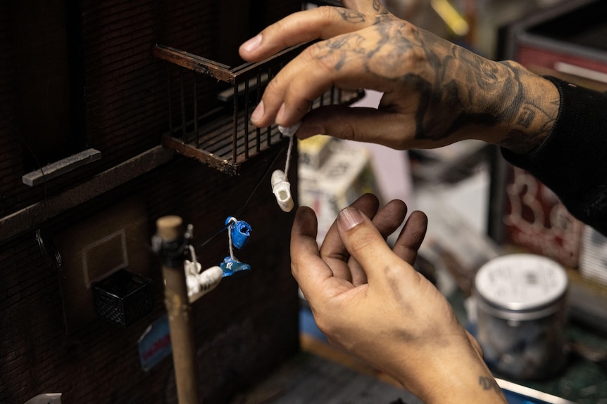 A pair of tattooed hands works on a miniature model of an urban street scene that includes sneakers hanging from a balcony
