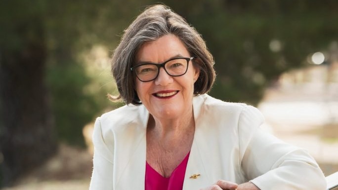 A close portrait of Cathy McGowan shows her in a white blazer sitting on a park bench. 