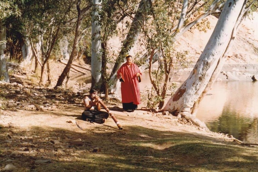 A boy sits on the ground playing a didgeridoo next to a man in red cloak and a river