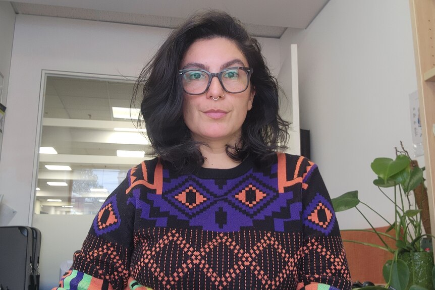 A woman sitting in an office with grey glasses and a blue, black and pink patterned jumper