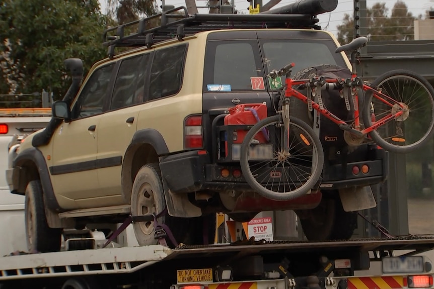 A taupe-coloured 4WD with a bike mounted on the back is towed on a trailer into the gates of the forensic centre.
