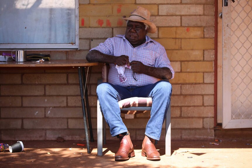 An Aboriginal man wearing jeans, checked shirt with rolled up sleeves, cowboy hat, sits on a chair in front of a brick wall.