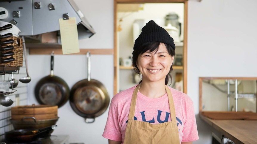Woman with black hair smiling into the camera standing in kitchen with apron on