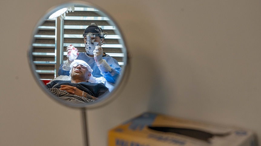 A doctor and patient are seen in the reflection of a mirror. The patient has bandages on his head