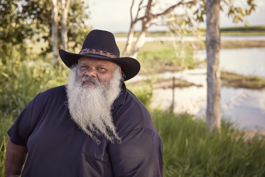 a man in a navy shirt with a long grey beard and a hat sits in front of a wetland.
