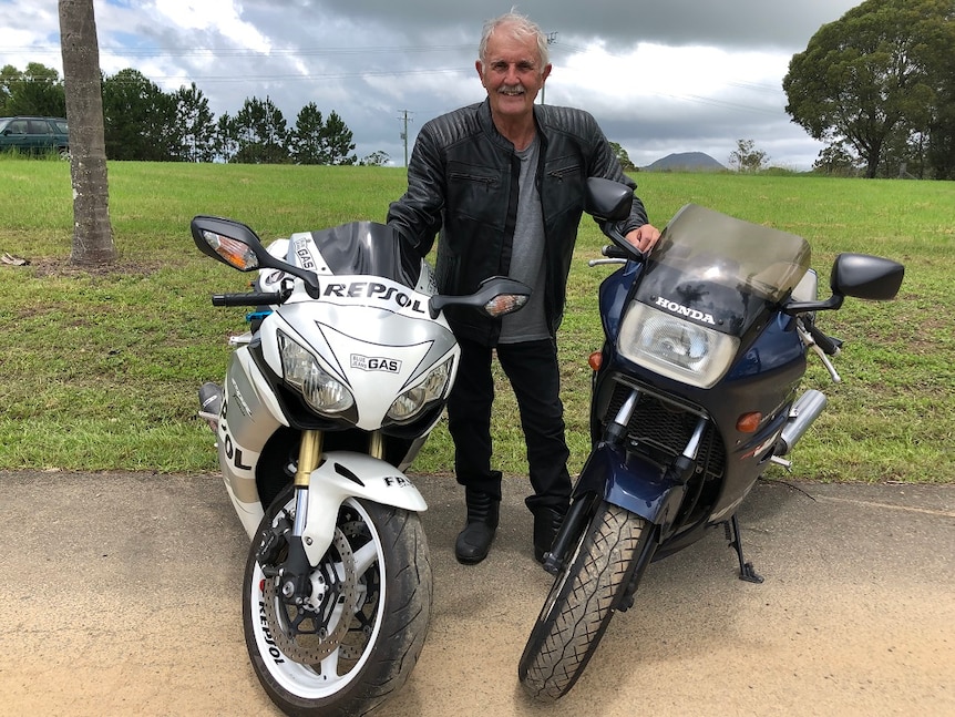 A man stands next to two motorcyles with green paddock behind him.