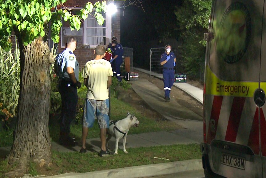 A nighttime scenes showing a police officer standing with a man and a white dog while an ambulance officer walks past. 