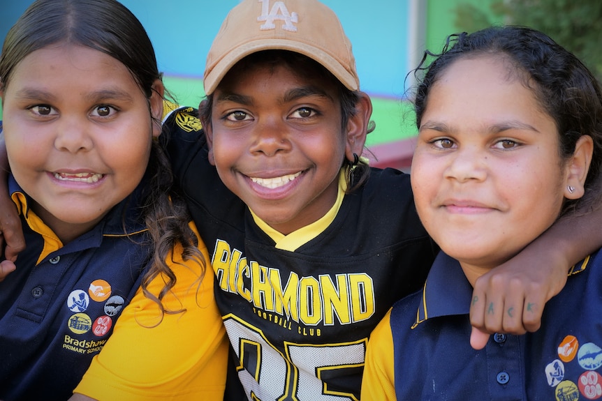 A close up of three Indigenous children, two girls, one boy in school uniform. Boy wears cap.