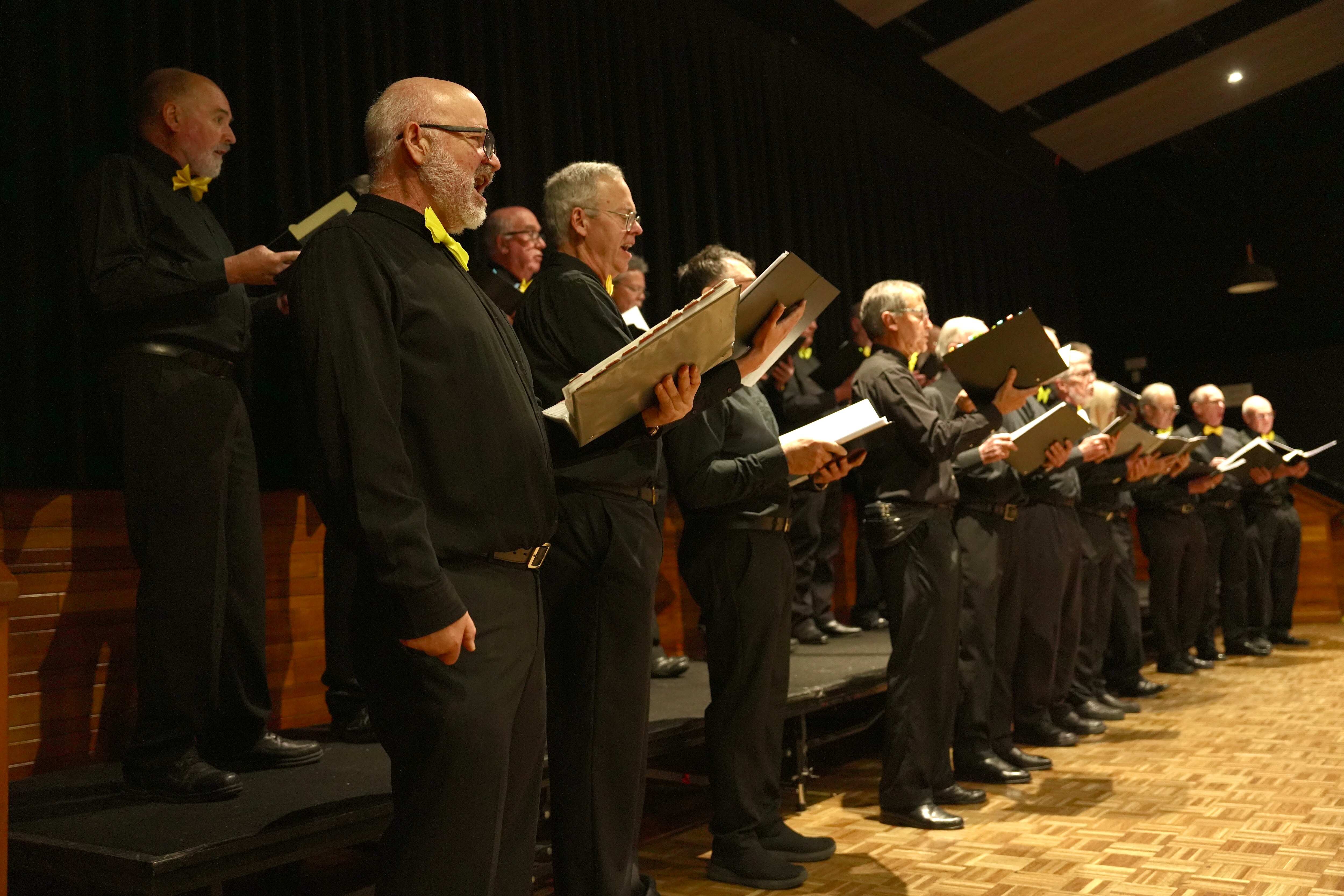 A male choir rehearses. There are two lines of men, with one line standing on a stop behind the others.