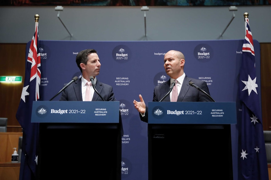 Two men in suits stand in front of podiums and banners marketing the 2021 budget, with Australian flags standing on either side.