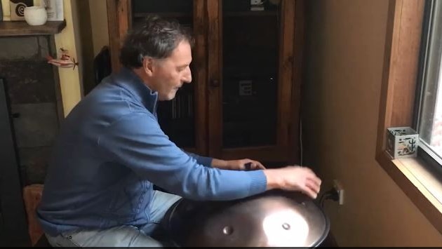 Man wearing long-sleeved blue jumper plays dome-shaped instrument with his right hand.