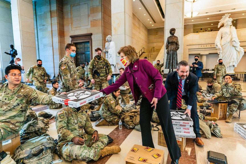 A woman wearing a mask and purple coat hands a pizza to a group of people in uniforms and wearing masks