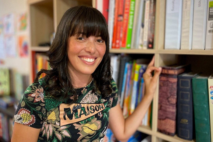 A young mother in a green patterned top stands next to a bookshelf full of books.