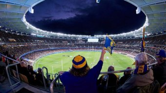 A fisheye shot of a full Perth Stadium at night, with a fan waving an Eagles flag in the foreground