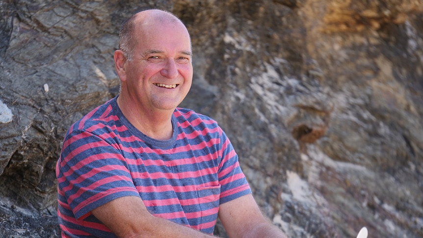 Program presenter Bill Wedgwood with his surfboard sitting on a rocky outcrop